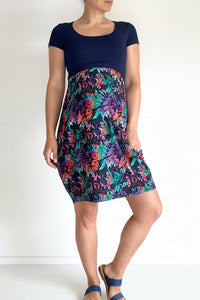 A woman modeling a short-sleeve dress with a solid navy blue bodice and a vibrant, multicolored floral print skirt featuring shades of pink, purple, teal, and orange.