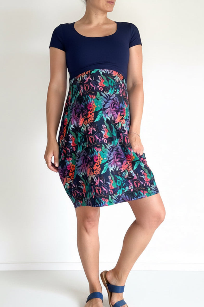 A woman modeling a short-sleeve dress with a solid navy blue bodice and a vibrant, multicolored floral print skirt featuring shades of pink, purple, teal, and orange.