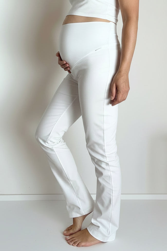 A pregnant woman wearing a matching white maternity set consisting of a cropped camisole top and high-waisted pants, standing barefoot on a light wood floor against a plain white wall.