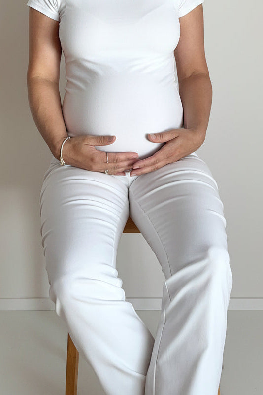 A pregnant woman sitting on a wooden stool, wearing a white ribbed camisole tank top and matching white pants against a plain off-white wall.