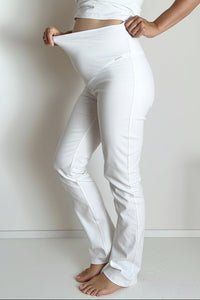 White maternity pants worn by a model, shown from the waist down as they stretch the elastic waistband with both hands.