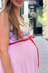 Woman in a pink maternity dress standing in an outdoor setting with a building in the background
