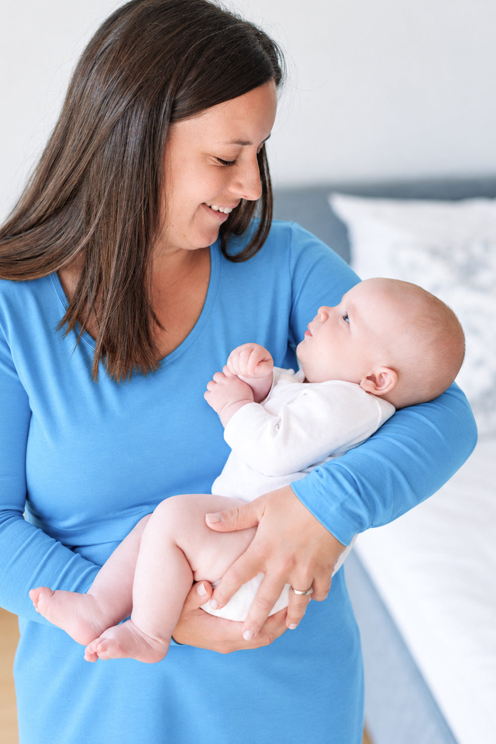 mamica v azurno modri spalni srajci za dojenje z dolgimi rokavi drži dojenčka v belem bodiju v spalnici z mehko osvetlitvijo.