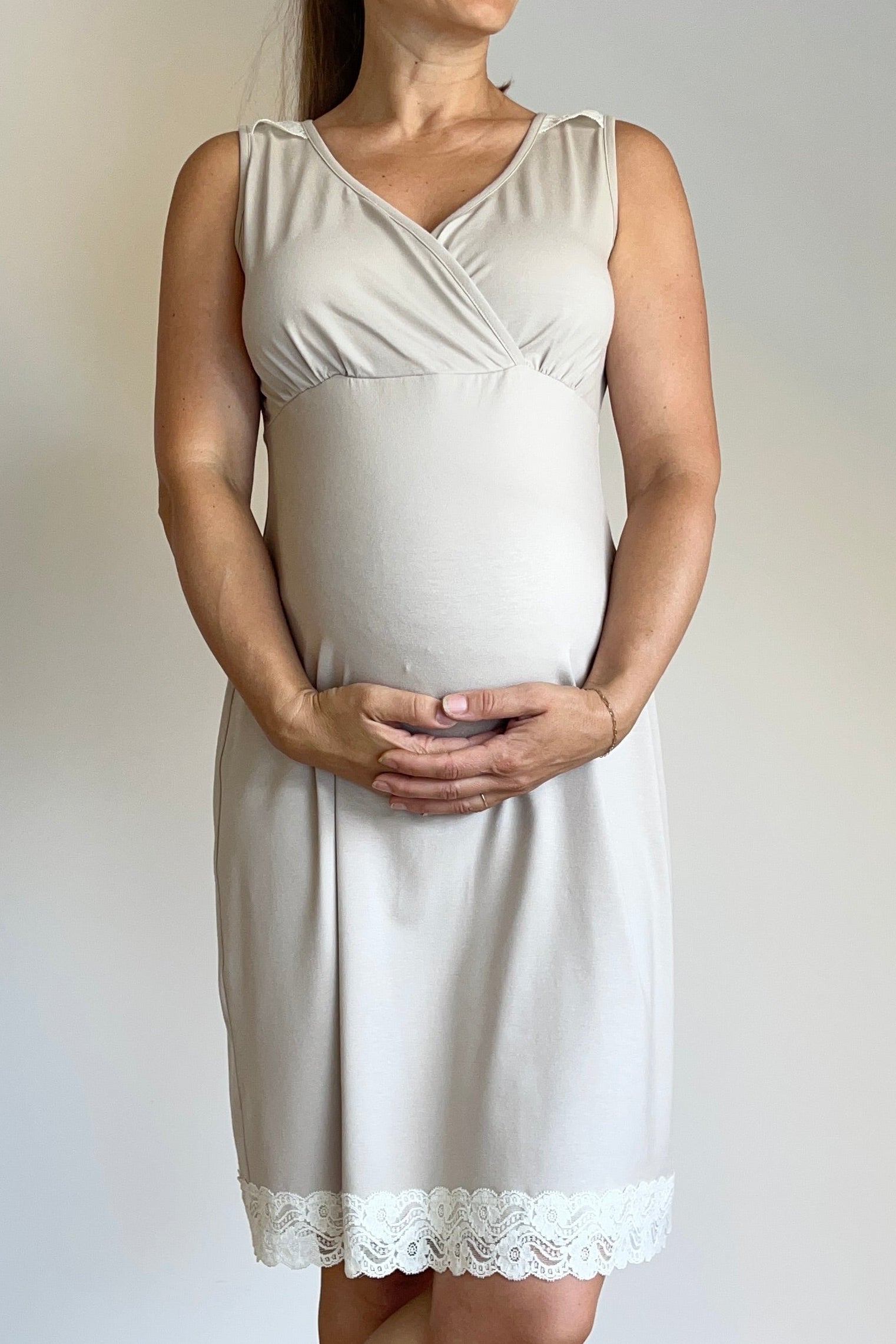 Woman wearing a white sleeveless dress with lace trim, standing against a plain white wall.
